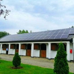 Tech-Systems24 sp. z o.o - Panele słoneczne na dachu budynku stajni z kolumnami, widoczne drzewa iglaste i osoba w tle.