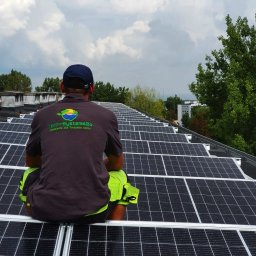 Tech-Systems24 sp. z o.o - Instalator siedzi na dachu pokrytym panelami fotowoltaicznymi, widok z tyłu, koszulka z logo Tech-Systems24, w tle budynki i drzewa pod pochmurnym niebem.