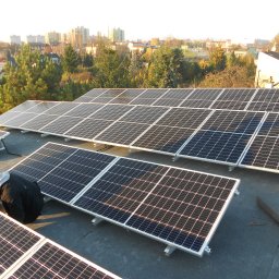 Tech-Systems24 sp. z o.o - Instalacja paneli słonecznych na płaskim dachu budynku z widokiem na miasto w tle, pracownik w czarnej kurtce pochylony nad panelami.