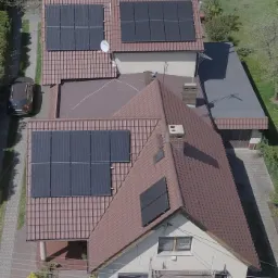 Widok z góry na domy z brązowymi dachami, na których zamontowane są panele fotowoltaiczne. Jeden z domów ma rusztowanie przy ścianie.