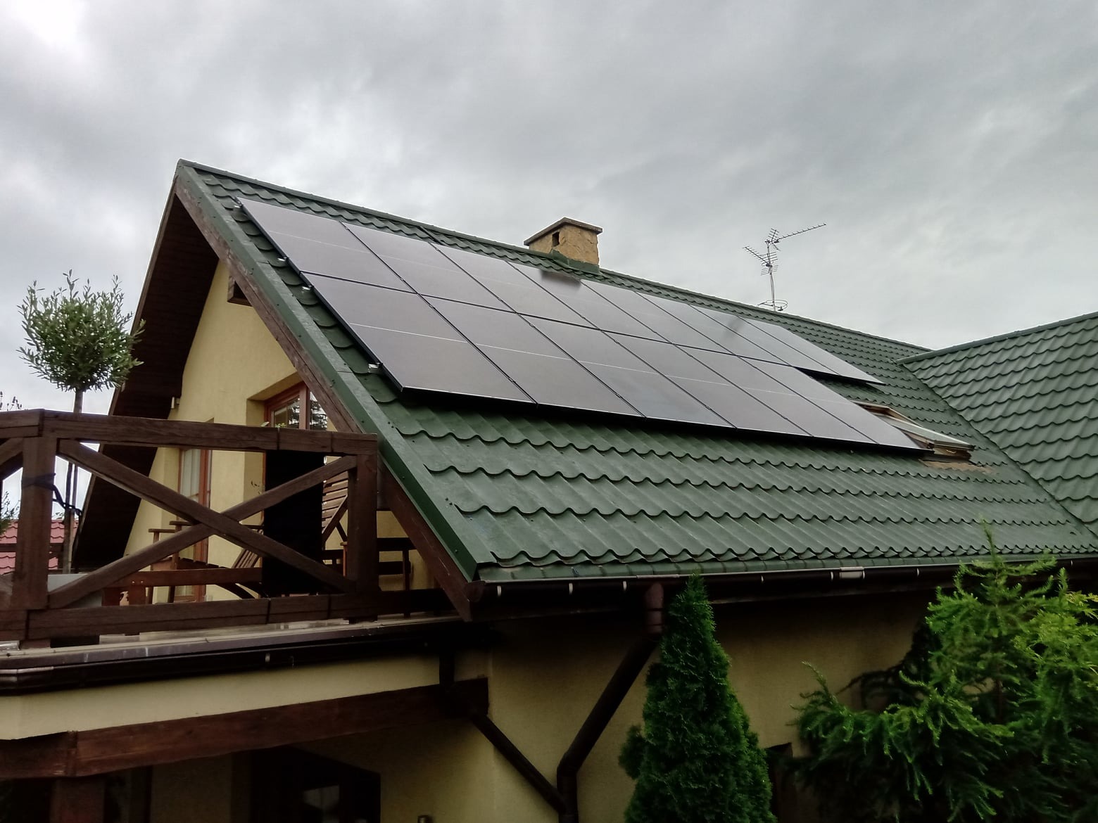 Panele słoneczne zamontowane na dachu domu z zieloną dachówką, widoczne z poziomu balkonu z drewnianą balustradą, na tle pochmurnego nieba.