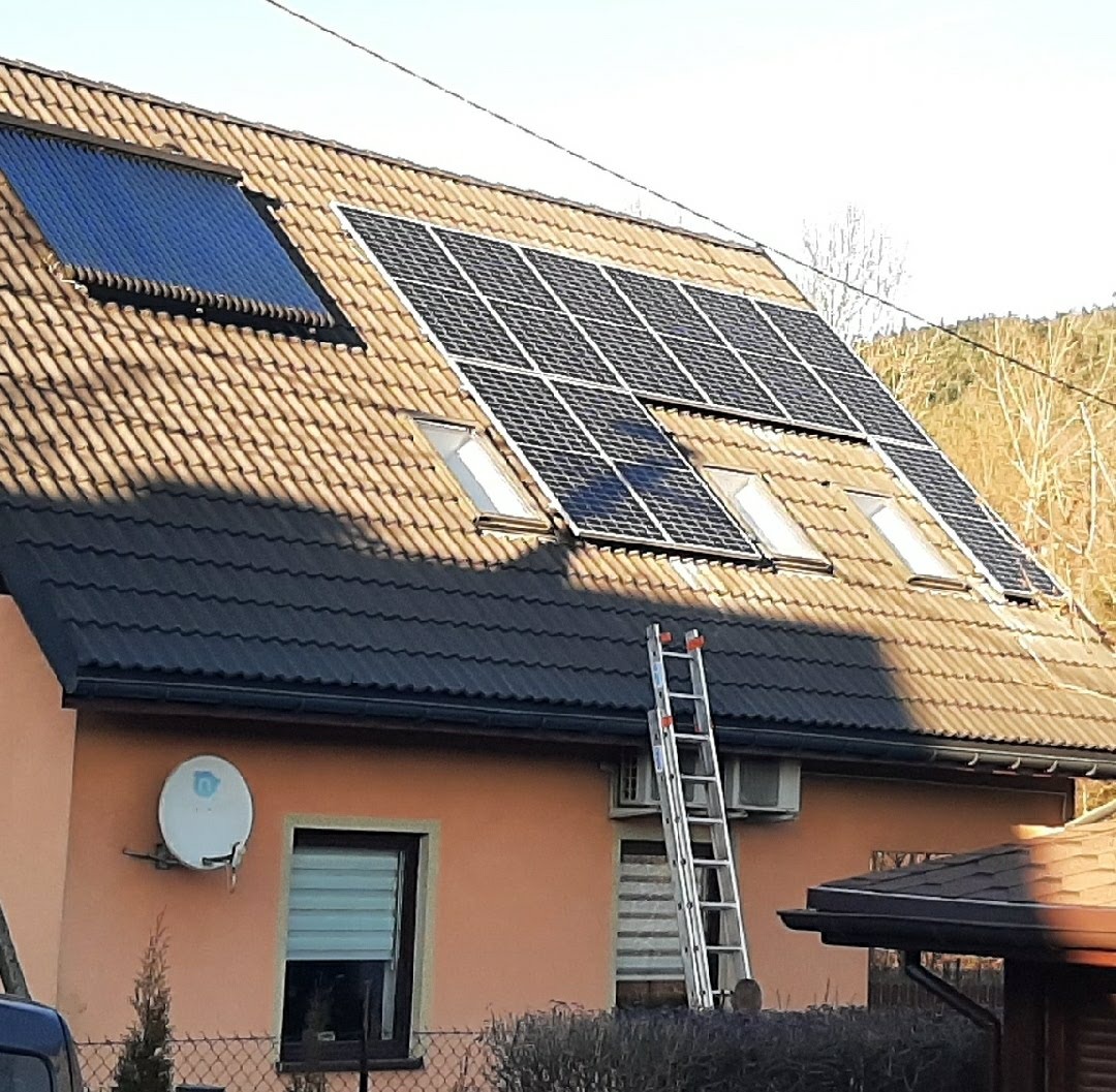 Dom z panelami słonecznymi i kolektorem słonecznym na dachu pokrytym brązową dachówką, drabina oparta o ścianę, antena satelitarna na elewacji.