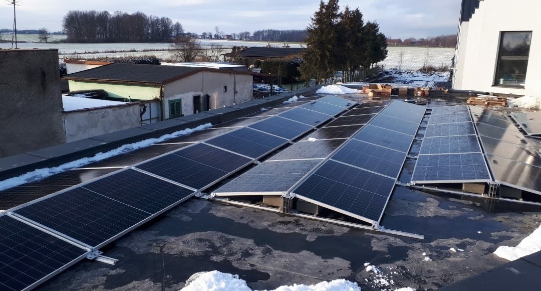 Panele słoneczne w trakcie montażu na płaskim dachu pokrytym papą, widoczne skrzynki i palety z elementami instalacji, resztki śniegu na dachu i w tle zabudowania gospodarcze.