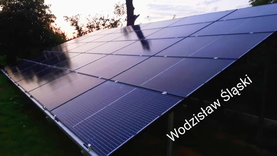 Instalacja paneli słonecznych na konstrukcji wsporczej, widok z dołu pod kątem, w tle drzewa i zachodzące słońce.