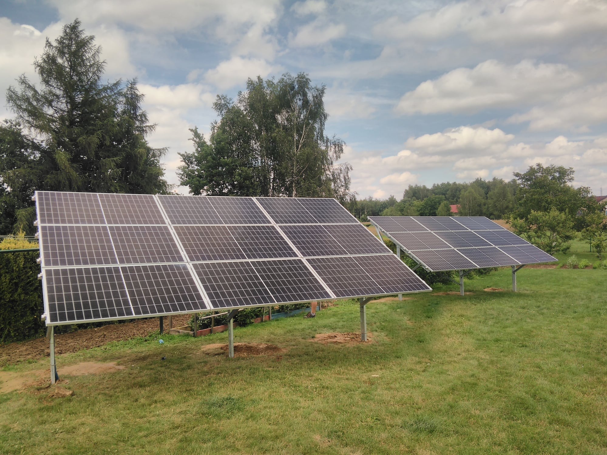 Dwa rzędy paneli słonecznych na metalowych podporach, zainstalowane na trawiastym terenie z drzewami i krzewami w tle pod błękitnym niebem z chmurami.