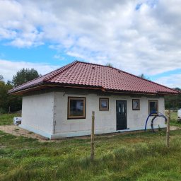 M&S-Bud - Niedokończony dom z czerwonym dachem z bloczków, okna i drzwi zamontowane, widoczne ocieplenie fundamentów, teren wokół ogrodzony siatką.