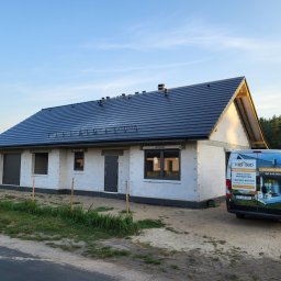 M&S-Bud - Dom w stanie surowym zamkniętym z ciemną dachówką, obok zaparkowany bus firmy budowlanej z reklamą gotowych domów.