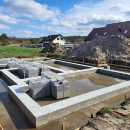 M&S-Bud - Zalane fundamenty domu jednorodzinnego z bloczków betonowych, widok z góry, w tle domy i hałdy ziemi, słoneczny dzień.