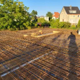 Zbrojenie płyty fundamentowej na drewnianym szalunku, widok z góry w słoneczny dzień, z widocznym cieniem osoby i zabudową w tle.