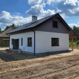 Nowoczesny dom jednorodzinny z dwukolorową elewacją (biały dół, szary góra) i ciemną dachówką, widoczny w słoneczny dzień na tle błękitnego nieba z chmurami, teren wokół domu jeszcze...