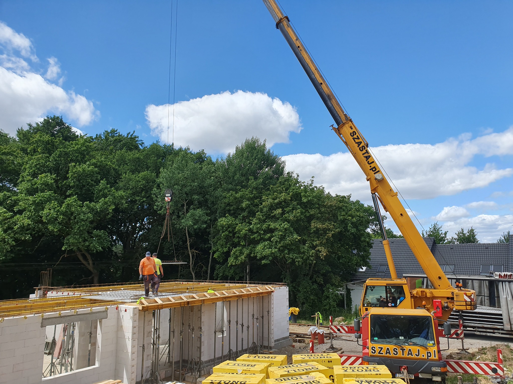 Budowa domu z bloczków Ytong: Dwóch robotników na stropie, żółty dźwig Szastaj.pl w tle, błękitne niebo z obłokami.
