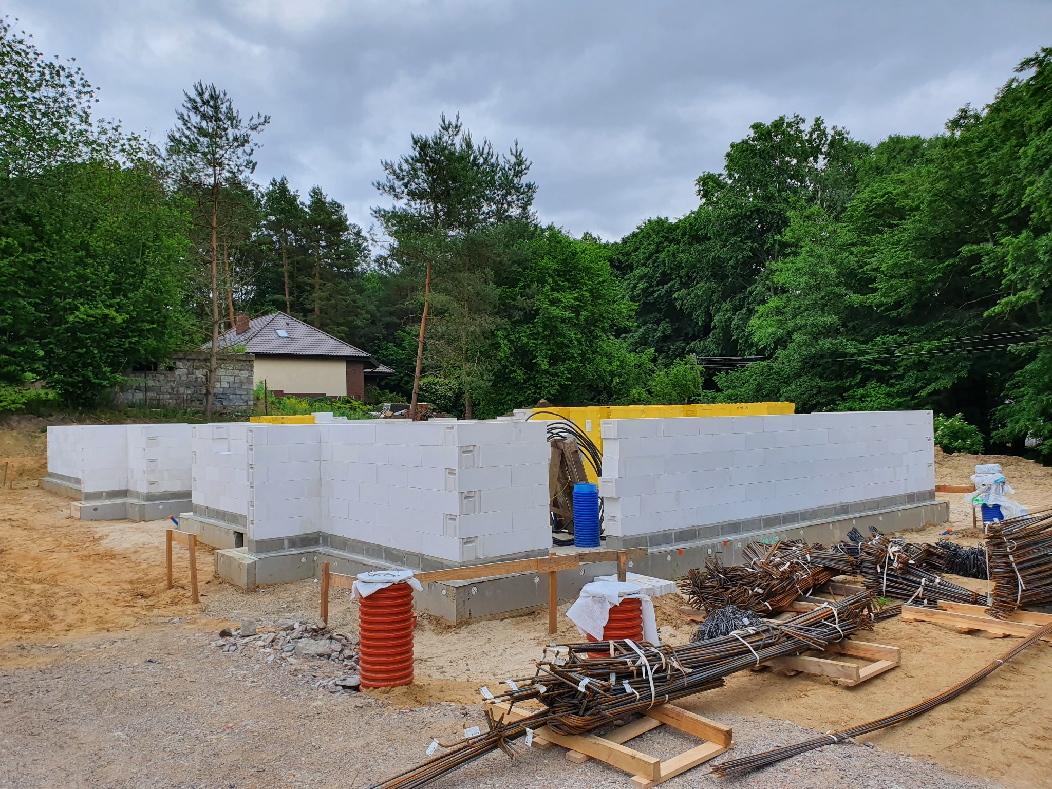 Budowa fundamentów domu z bloczków betonowych, widoczne zbrojenia, pomarańczowe rury i żółte elementy izolacyjne, w tle zieleń i fragment domu.