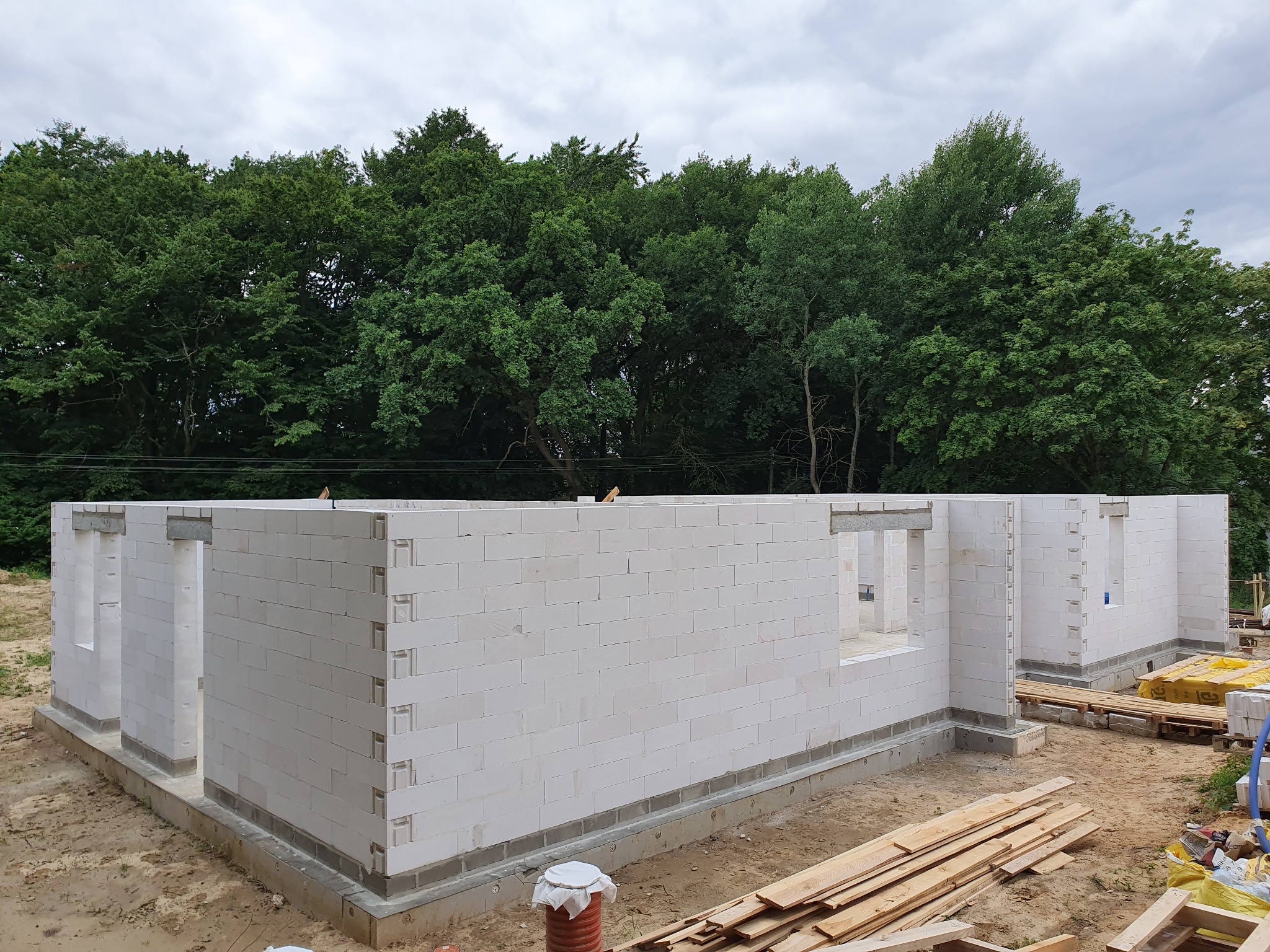 Wznoszone ściany parterowe domu z białych bloczków, widoczne otwory okienne, w tle zieleń drzew, na ziemi deski i palety z materiałami budowlanymi.