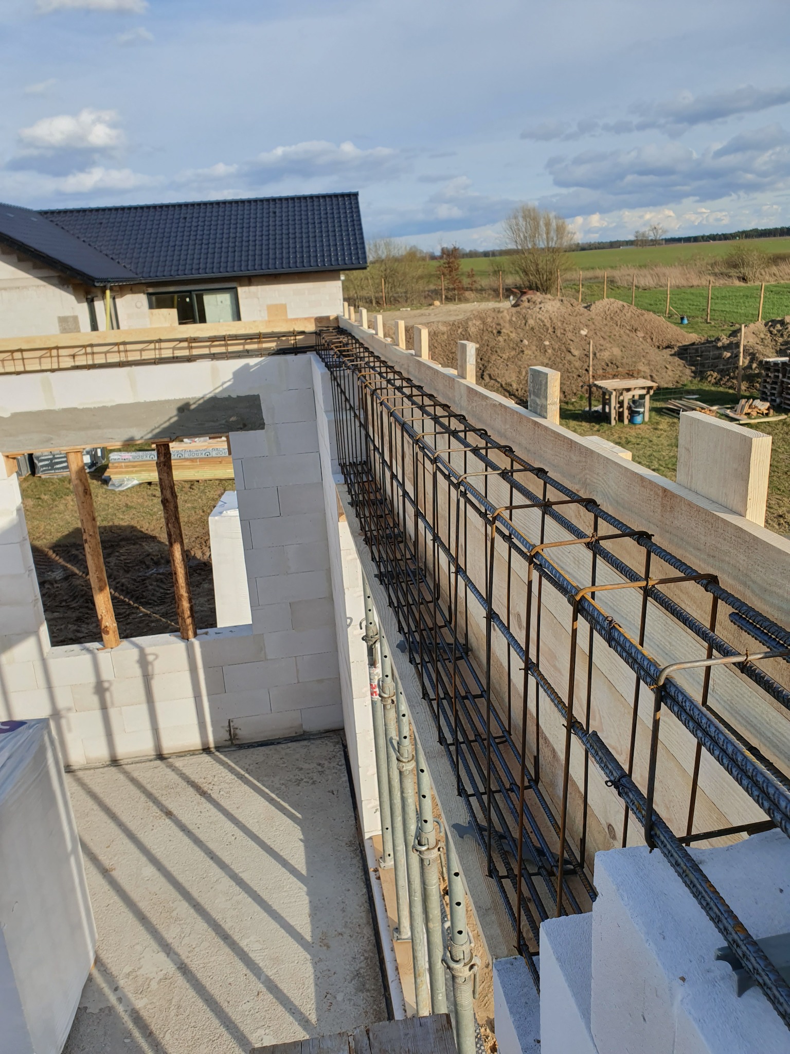 Zbrojenie wieńca na murze z betonu komórkowego w trakcie budowy domu jednorodzinnego, widok z góry na szalunek i stalowe pręty, w tle krajobraz z polami i lasem.