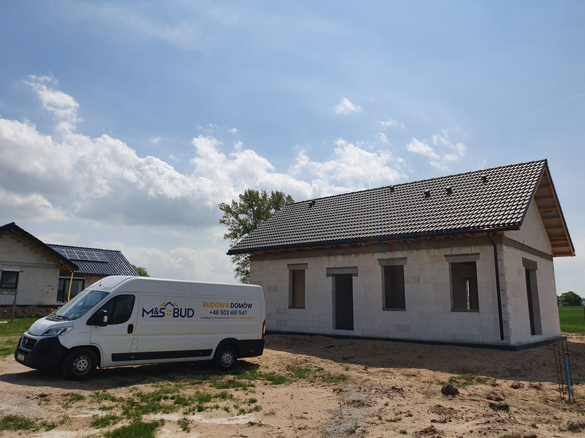 Wznoszony dom z betonu komórkowego z ciemną dachówką, na tle błękitnego nieba z obłokami, obok biały bus firmy budowlanej z widocznym logo i numerem telefonu.