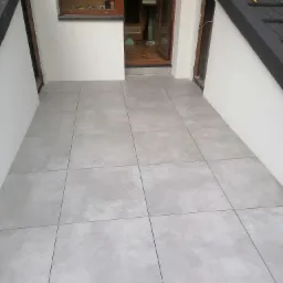 Wyłożony szarymi płytkami taras z widocznym narzędziem do silikonu na dachu, częściowo wykończony, z otwartymi drzwiami wejściowymi.