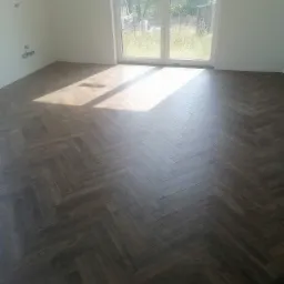 Ułożona parkiet w jodełkę z ciemnego drewna w jasnym pokoju, widoczne instalacje elektryczne i duże okno balkonowe z widokiem na ogród.