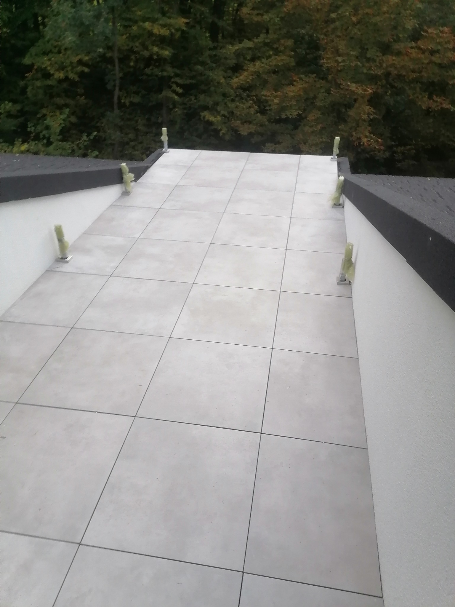 Jasnoszary taras z płytek ceramicznych w trakcie budowy, widoczne elementy montażowe zabezpieczone folią, tło stanowią drzewa.