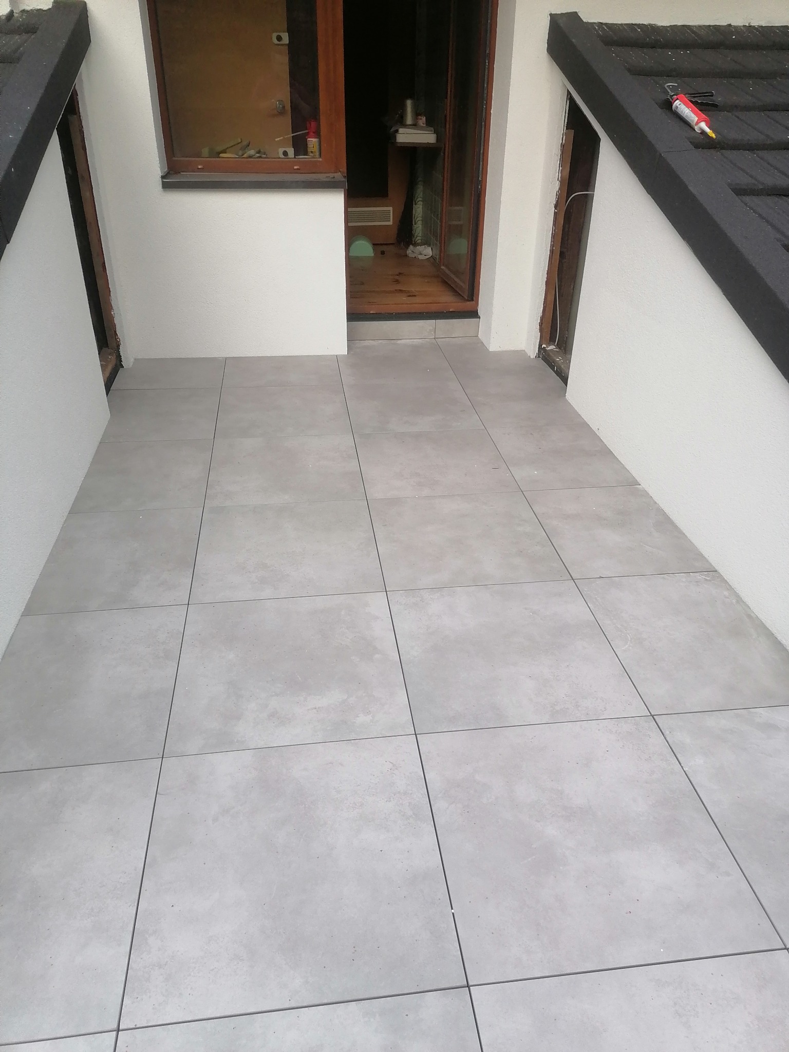 Wyłożony szarymi płytkami taras z widocznym narzędziem do silikonu na dachu, częściowo wykończony, z otwartymi drzwiami wejściowymi.