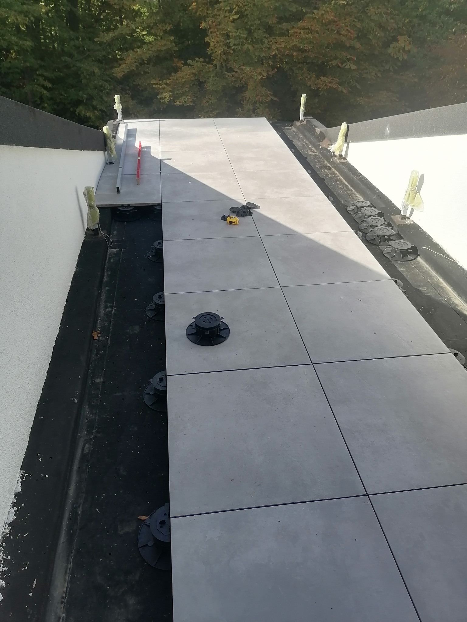 Montaż szarego tarasu z płyt na regulowanych wspornikach, widoczne poziomice i narzędzia, czarna hydroizolacja pod spodem.