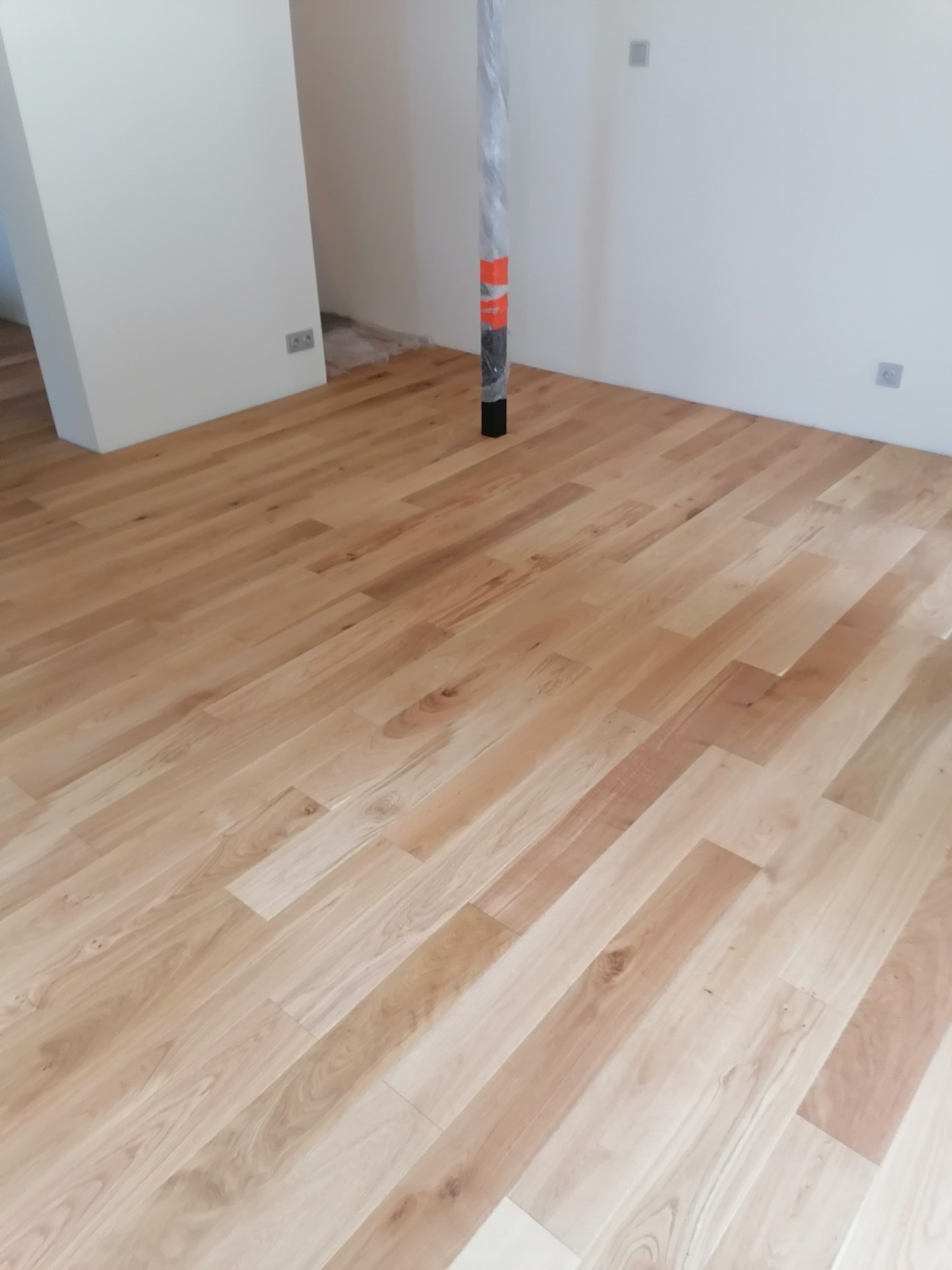 Jasne, drewniane deski podłogowe ułożone w nowym pomieszczeniu z białymi ścianami i słupem zabezpieczonym folią ochronną.