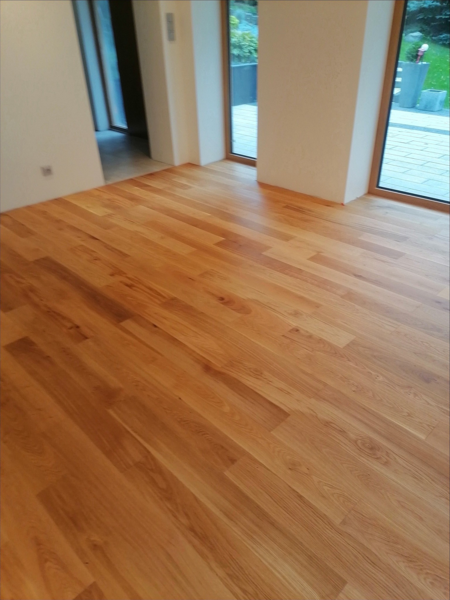 Nowo położona, lakierowana podłoga z jasnego drewna w pomieszczeniu z dużymi oknami wychodzącymi na ogród. Widoczne ściany w kolorze złamanej bieli i drewniane ramy okienne.