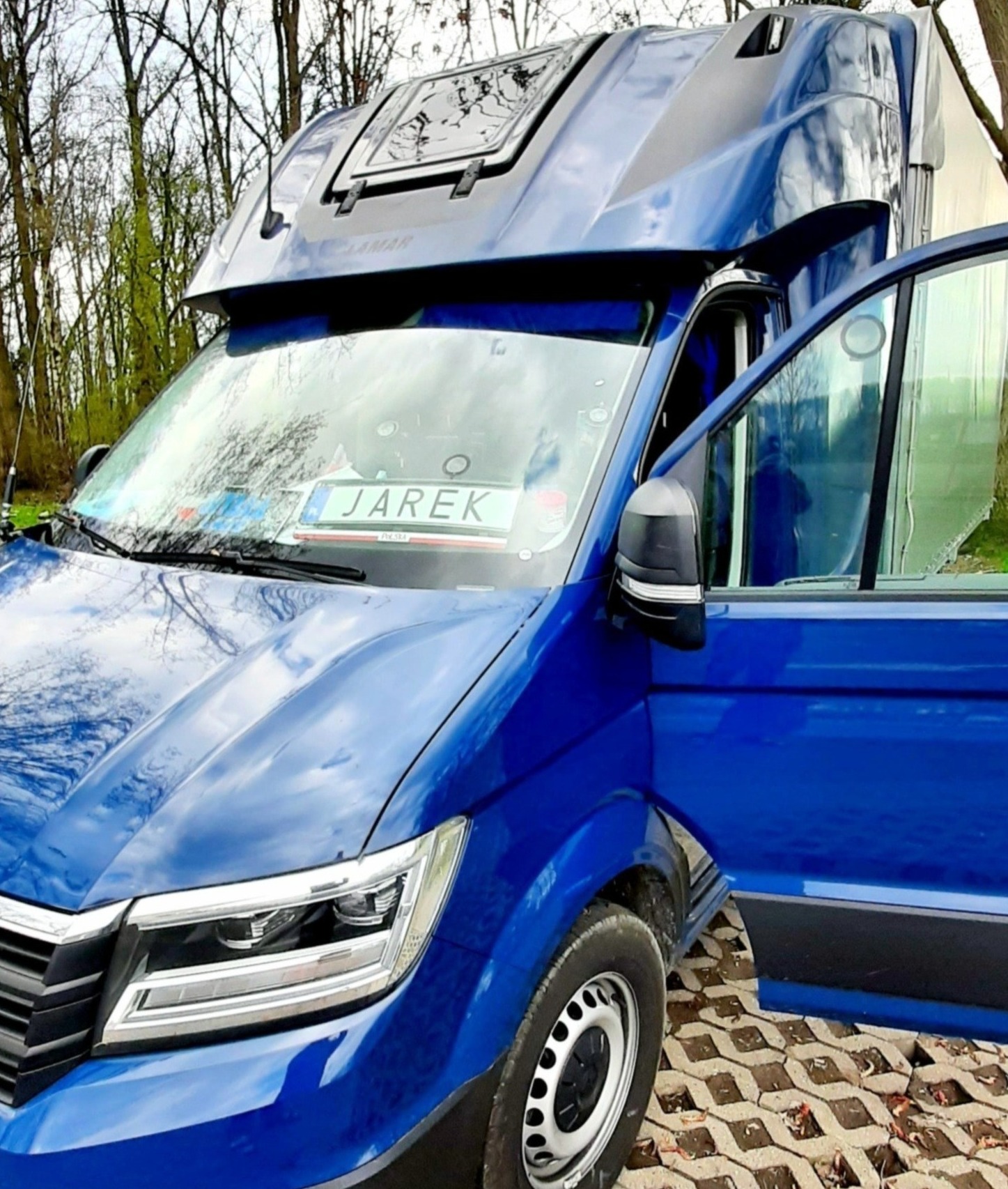 Niebieski bus z otwartymi drzwiami pasażera, widoczna tablica rejestracyjna z imieniem 'JAREK' na przedniej szybie, na tle drzew i częściowo widocznego białego pojazdu.