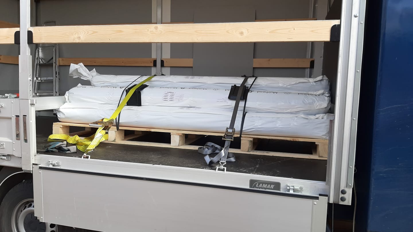 Ładunek rolek materiału na palecie, zabezpieczony pasami transportowymi w przestrzeni ładunkowej busa z drewnianą konstrukcją mocującą.