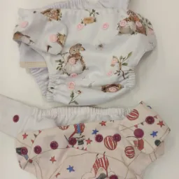 Dwa pieluszki wielorazowe z różnymi wzorami leżące na białym tle. Jedna z motywem królików i kwiatów, druga z motywem cyrkowym i gwiazdami, obie zapinane na zatrzaski.