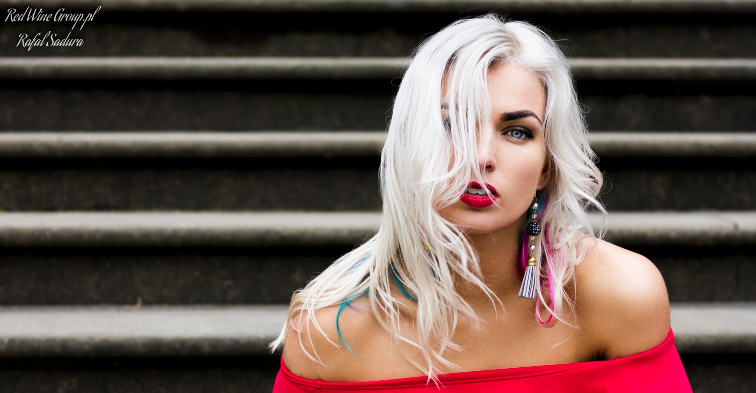 Portret kobiety z platynowymi włosami i czerwonymi ustami, w czerwonej bluzce na tle betonowych schodów, z widocznym kolczykiem.