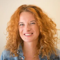 Uśmiechnięta kobieta z burzą rudych, kręconych włosów, ubrana w jeansową kurtkę, na neutralnym tle.