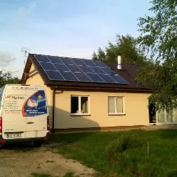 Dom jednorodzinny z panelami słonecznymi na dachu, obok zaparkowany biały bus firmy Hymon z otwartymi drzwiami, widoczny trawnik i drzewa.