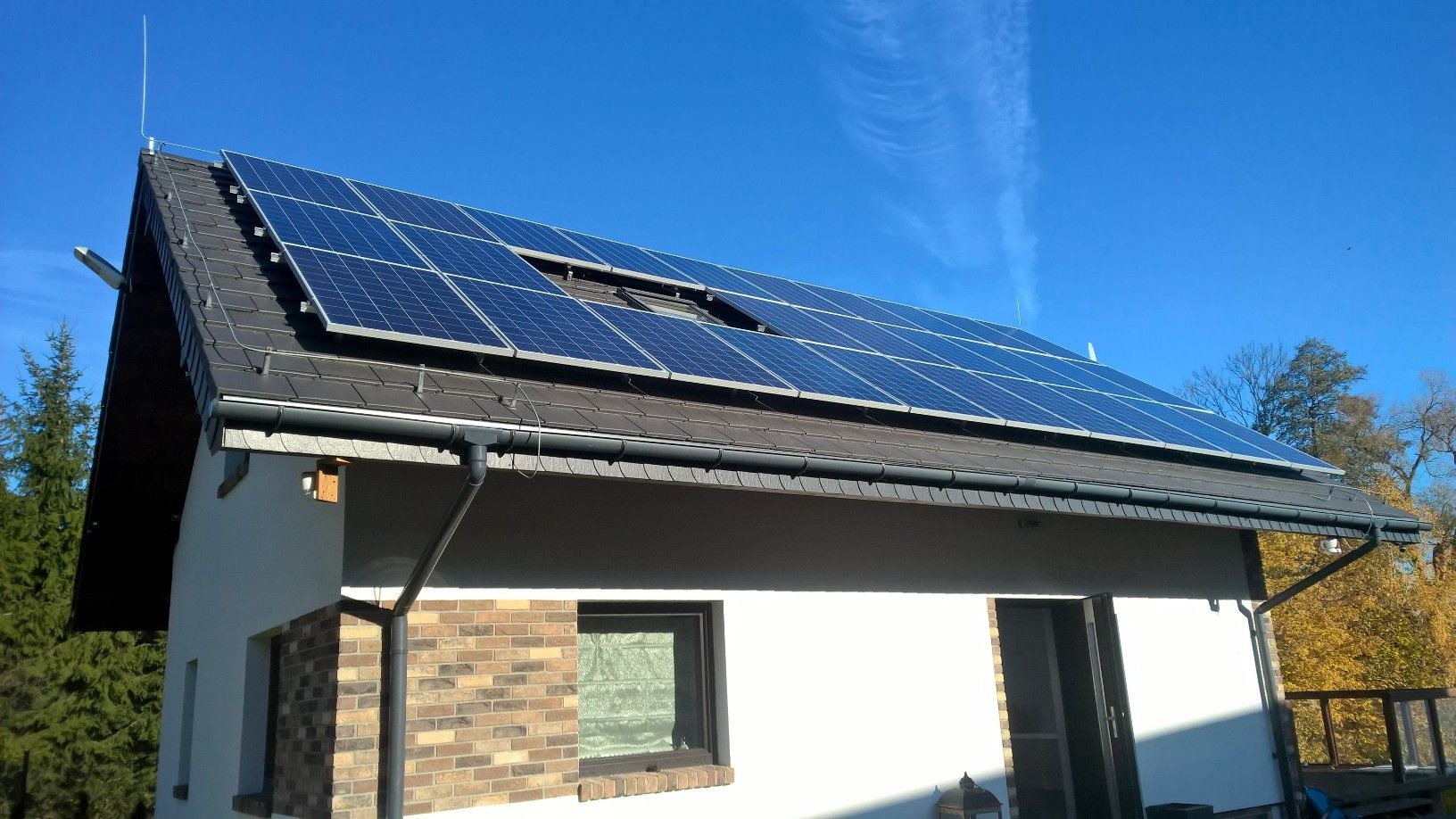 Instalacja paneli słonecznych na dachu dwuspadowym domu jednorodzinnego z brązową dachówką, widok z dołu na tle błękitnego nieba.