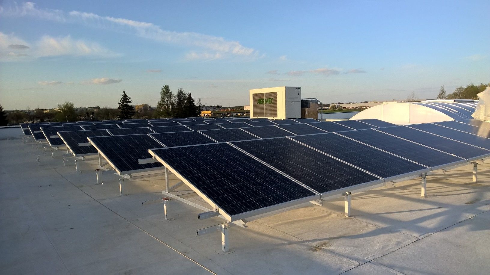 Rząd paneli słonecznych na płaskim dachu budynku z widoczną jednostką klimatyzacyjną AERMEC w tle, pod błękitnym niebem z lekkimi chmurami.