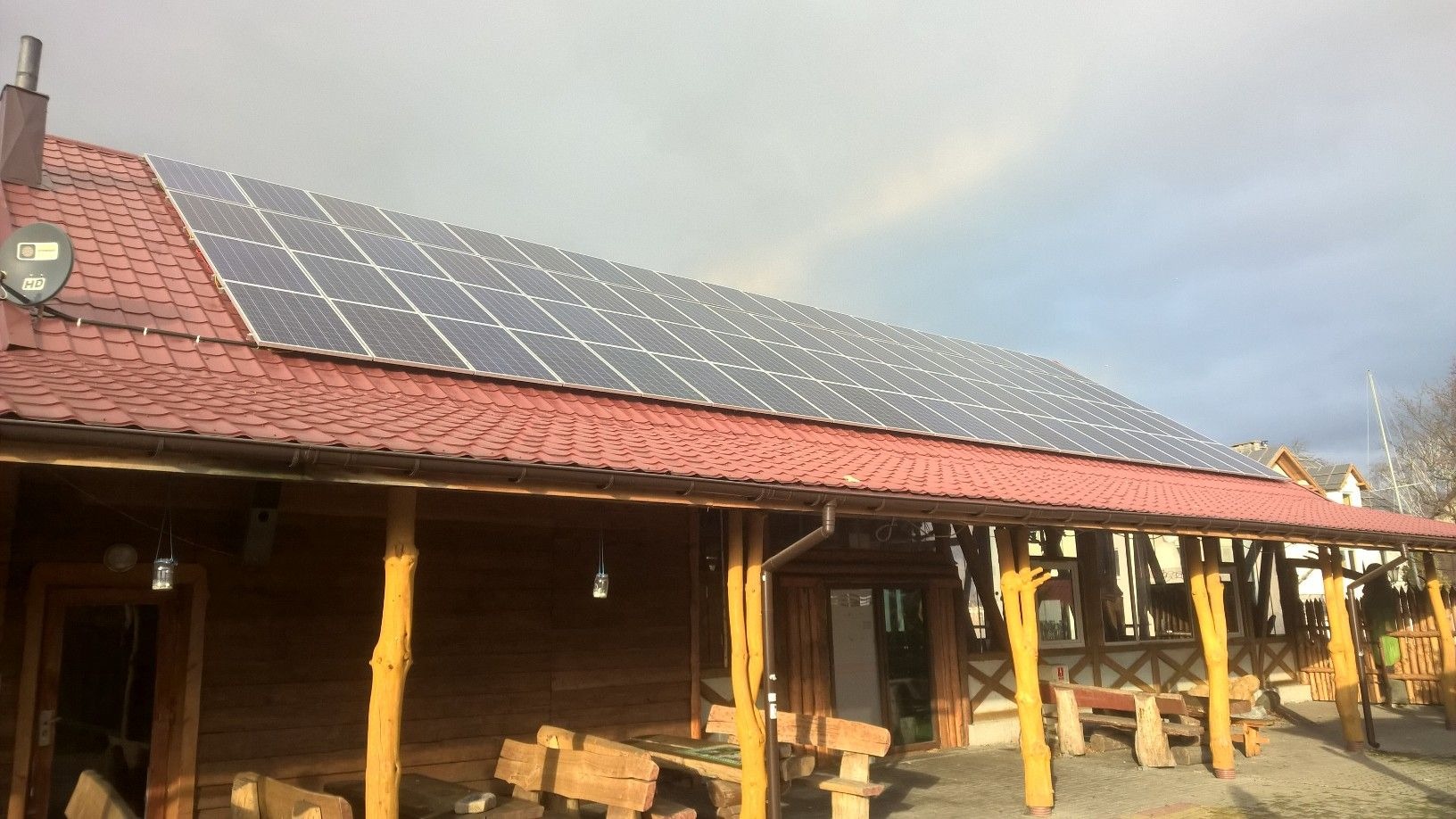 Instalacja paneli słonecznych na dachu drewnianej karczmy z czerwoną dachówką, widoczna antena satelitarna.