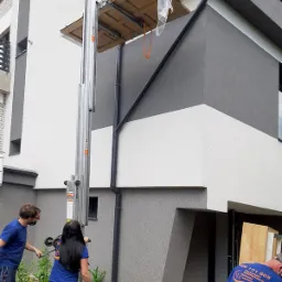 Winda meblowa transportująca drzwi na platformie, pracownicy w niebieskich koszulkach obsługują urządzenie przy szaro-białym budynku.