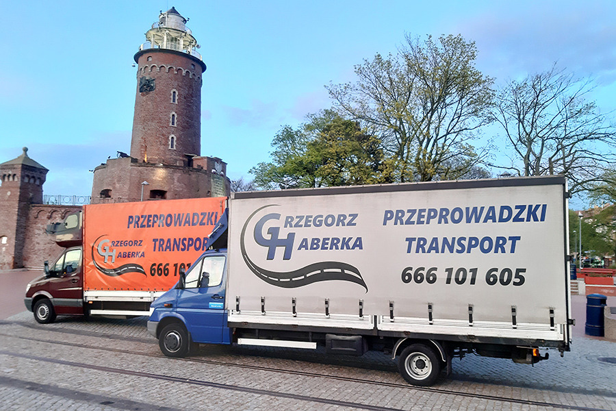 Dwa busy transportowe firmy Grzegorz Haberka zaparkowane na tle ceglanej latarni morskiej w Świnoujściu, jeden pomarańczowy, drugi niebieski, oba z logo firmy i numerem telefonu.