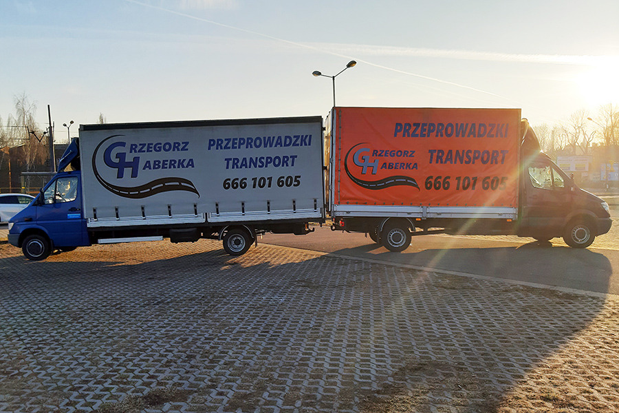Dwa samochody dostawcze z naczepami, z reklamą firmy transportowej Grzegorz Aberka, zaparkowane na placu z ażurowej kostki brukowej w słoneczny dzień.