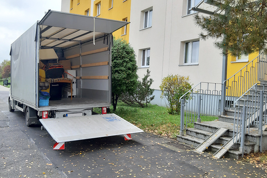 Otwarta paka busa wypełniona meblami i kartonami na tle bloku mieszkalnego z rampą dla wózków inwalidzkich.