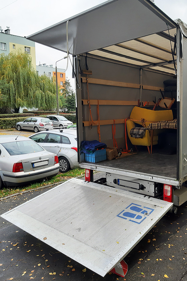 Otwarta rampa załadunkowa busa z widocznym wnętrzem: żółty fotel, kartony i niebieskie pudło; w tle zaparkowane samochody i budynek mieszkalny.