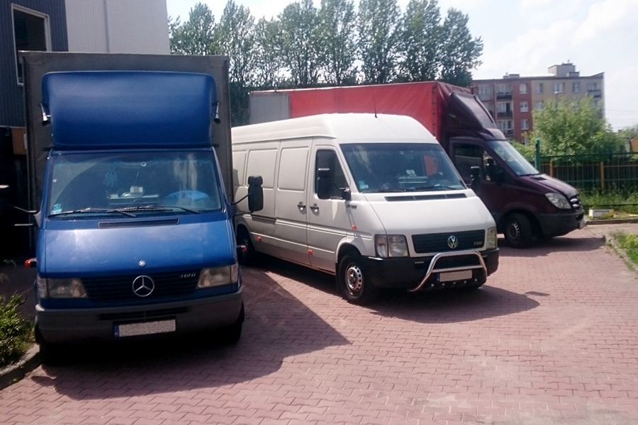 Trzy busy transportowe różnych marek (Mercedes, Volkswagen, Mercedes) zaparkowane na brukowanym placu przed budynkiem mieszkalnym.