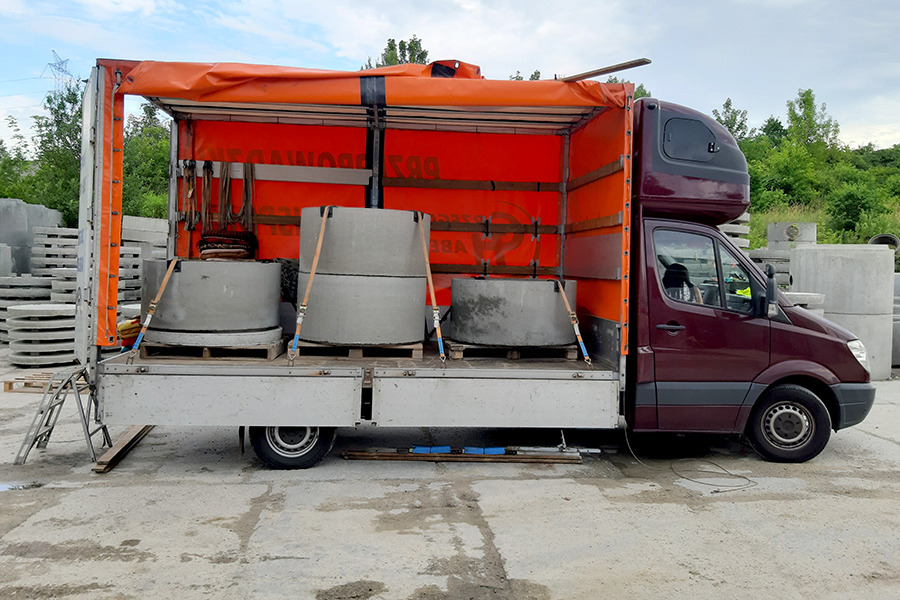Ciemnoczerwony bus z otwartą przestrzenią ładunkową, zabezpieczony pomarańczową plandeką, przewozi betonowe kręgi na paletach, zabezpieczone pasami transportowymi.
