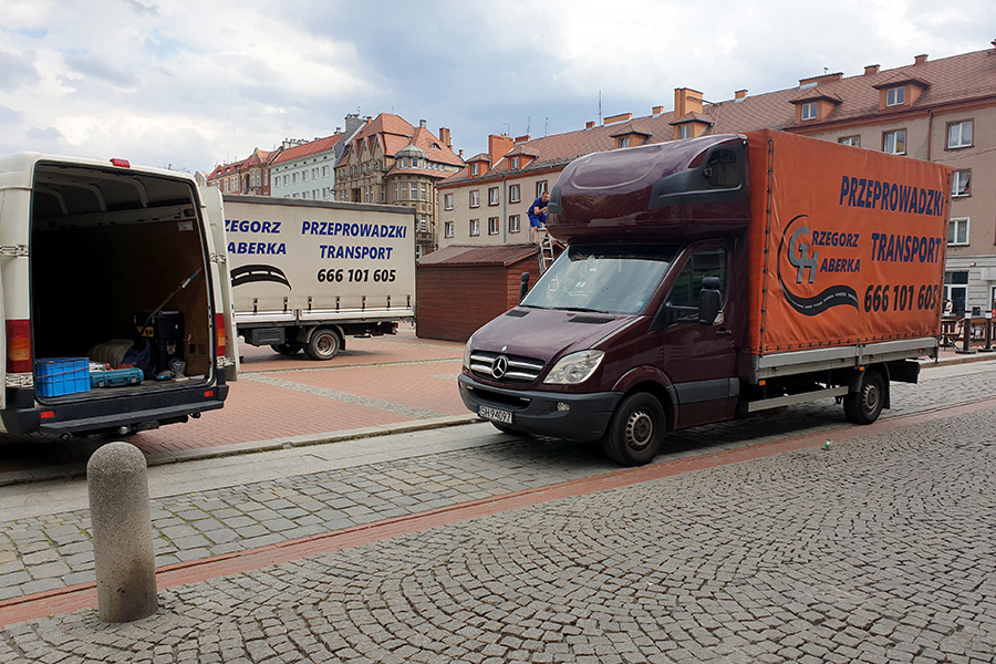 Trzy busy transportowe, w tym jeden z otwartym bagażnikiem z narzędziami, stojące na brukowanym placu miejskim z historyczną zabudową w tle. Na dachu jednego z busów stoi osoba.