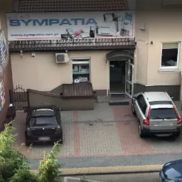 Fasada budynku z szyldem 'sklep SYMPATIA' prezentującym telefony i akcesoria, z zaparkowanymi samochodami na brukowanym podjeździe.