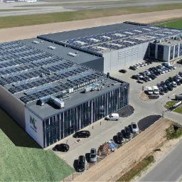 Widok z lotu ptaka na duży budynek przemysłowy z panelami słonecznymi na dachu i parkingiem pełnym samochodów.