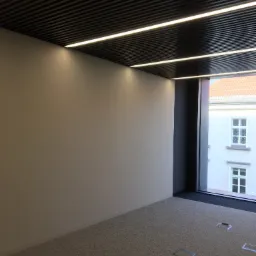 Wnętrze biurowe w Świdniku z wykończoną ścianą, widok na sufit z podwieszanymi panelami i oświetleniem liniowym, oraz okno z widokiem na budynek.