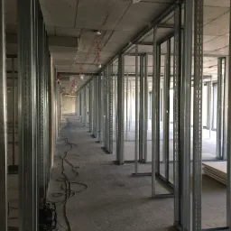 Stalowe profile konstrukcyjne tworzące szkielet ścian działowych w surowym wnętrzu, widoczne przewody elektryczne i lampy w folii ochronnej.