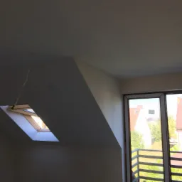 Poddasze po remoncie z oknem dachowym i balkonem, widoczne gładkie ściany i sufit, przewód zwisający z sufitu.
