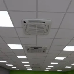 Kasetonowy sufit podwieszany z wbudowanymi panelami LED i dwoma klimatyzatorami kasetonowymi, widok z dołu w pomieszczeniu z zieloną ścianą.