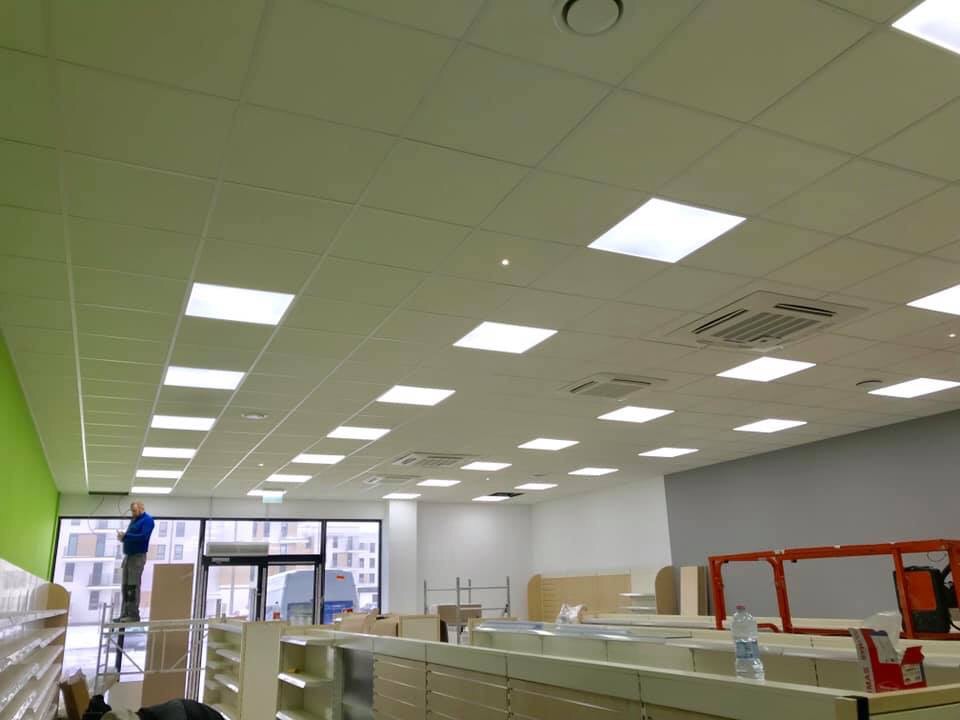 Montaż sufitu kasetonowego z wbudowanym oświetleniem w lokalu handlowym, widoczny pracownik na rusztowaniu i elementy wyposażenia sklepu.